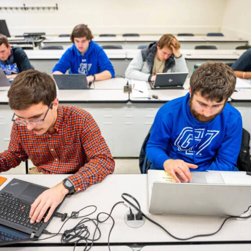 A classroom setting with students seated at long desks, each working on laptops. The students appear focused, with some typing, using a mouse, or reading. The room has a tiered layout with white walls and built-in outlets on the desks.